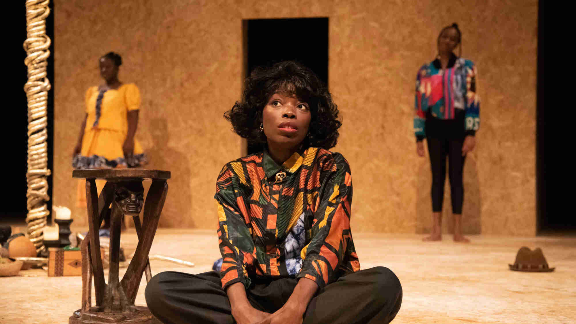 A female actor sat cross legged on stage. Two other actors stand in from of the wooden back wall behind her.