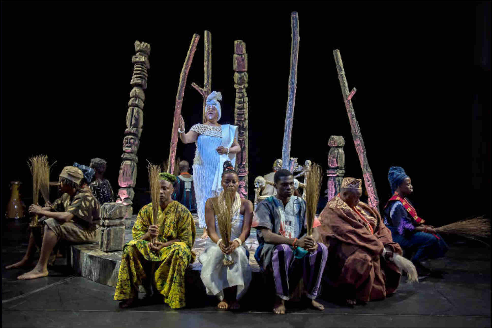 Performance of Duchess of Malfi. A row of performers sat on a stage with a stood female performer behind them. The stage is also covered in trees. All performers are dressed in traditional African Clothing